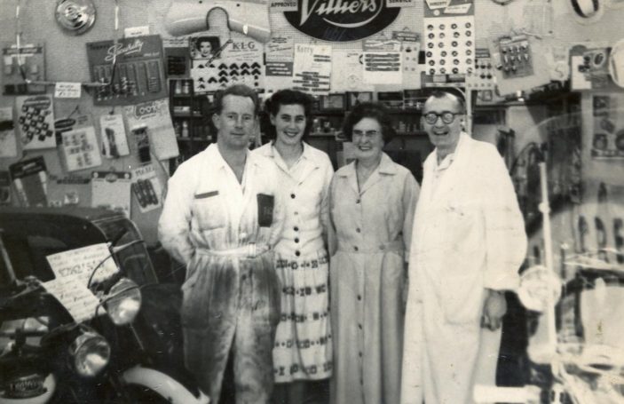 A black and white photograph of a shop interior showing four adults (two women and two men) standing in a line. Both men are wearing overalls, one pair very dirty, the other clean. The two women wear dresses. The walls behind them are covered with shop stock, mostly on cards.